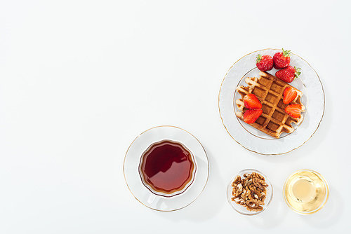 top view of waffle with strawberries on plate near cup with tea, bowls with honey and nuts on white