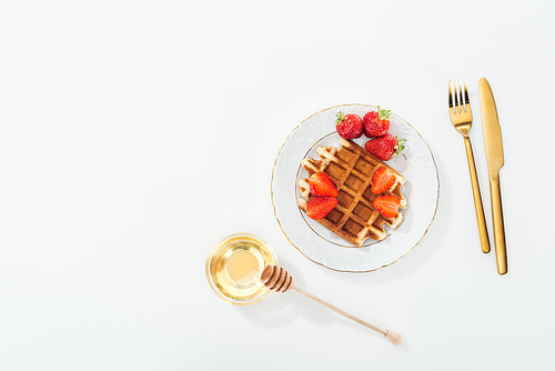top view of waffle with strawberries on plate near bowl with honey and wooden dipper, and cutlery on white
