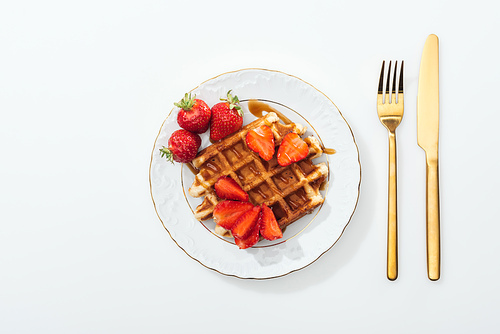 top view of plate with waffle and strawberries near fork and knife on white