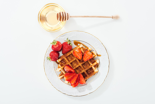 top view of bowl with honey and wooden dipper near plate with waffle and strawberries on white