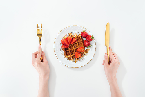 cropped view of woman holding fork and knife near plate with waffle and strawberries on white