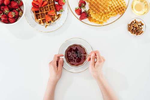 cropped view of woman holding teaspoon and cup of tea near plates with strawberries and waffles on white