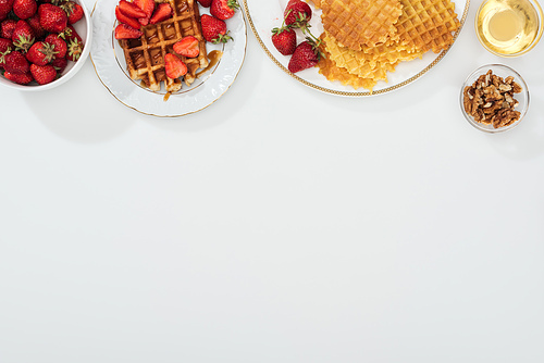top view of waffles and strawberries on plated near bowls with honey and nuts on white