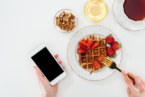 cropped view of woman eating breakfast and holding smartphone on white