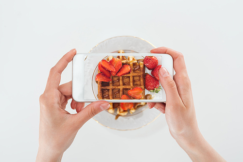 cropped view o woman taking picture of tasty waffle with strawberries on plate isolated on white