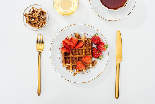 top view of served breakfast with waffle and strawberries on plate, honey and nits in bowls, cup of tea near fork and knife on white
