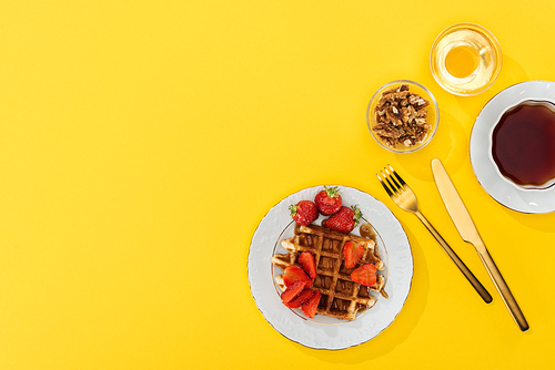top view of waffle and strawberries, nuts, honey and tea near cutlery on yellow