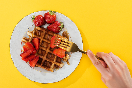 cropped view of woman taking strawberry from plate with waffle on yellow