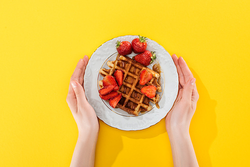 cropped view of woman holding plate with waffle and strawberries on yellow