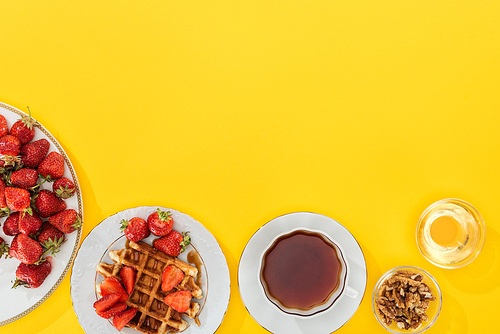 top view of plates with strawberries and waffles with cup of tea and bowl with nuts and honey on yellow