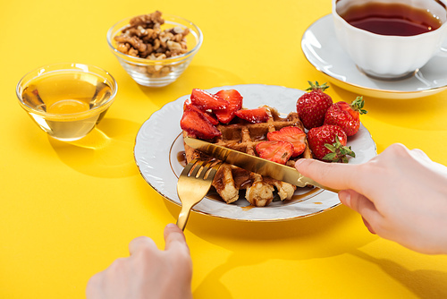 cropped view of woman cutting waffles on plate near bowl and cup with tea on yellow background