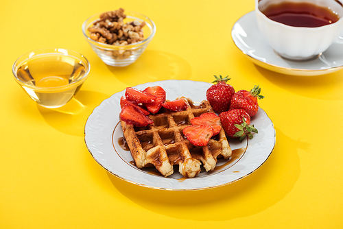 served breakfast with waffle and strawberries on plated, honey, nuts and tea on yellow background