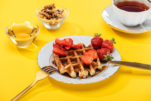 served breakfast with waffle and strawberries on plated near cutlery, honey, nuts and tea on yellow background