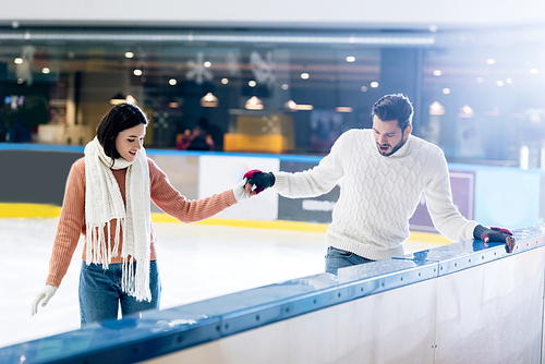 cheerful young woman teaching man to skate on a rink while holding hands