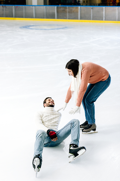 positive young couple skating together while man falling on rink