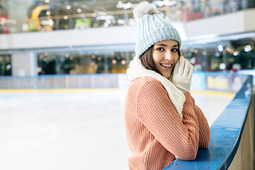 positive girl in sweater, scarf, gloves and hat standing on skating rink