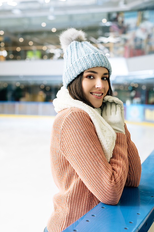 beautiful smiling girl in sweater, scarf, gloves and hat standing on skating rink