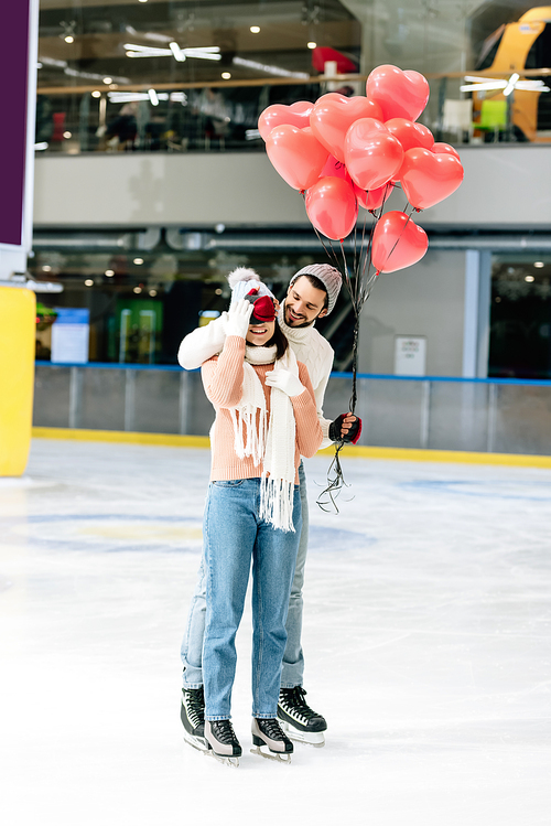 man with red heart shaped balloons closing eyes to girlfriend on skating rink on valentines day