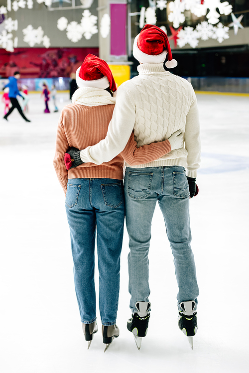 back view of happy couple in santa hats hugging on skating rink