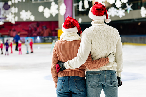 back view of young couple in santa hats hugging on skating rink at christmastime