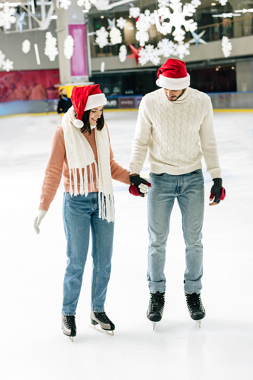 happy couple in santa hats holding hands while skating on rink