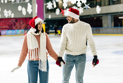 beautiful happy couple in santa hats holding hands at christmastime on skating rink