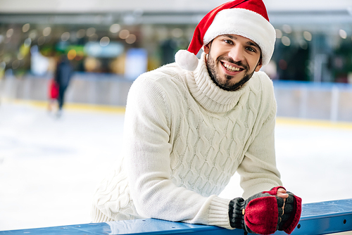 bearded smiling man in sweater and santa hat on skating rink