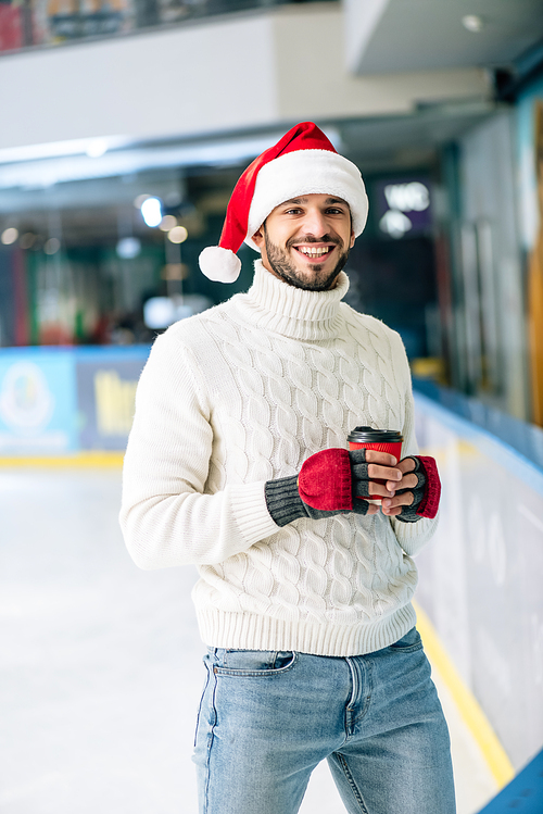 handsome cheerful man in sweater and santa hat holding coffee to go on skating rink