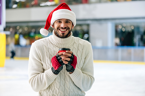 handsome cheerful man in santa hat holding coffee to go on skating rink