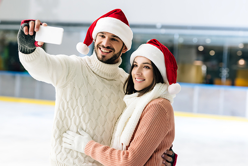 smiling couple in santa hats taking selfie on smartphone on skating rink at christmastime