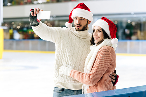 cheerful couple in santa hats taking selfie on smartphone on skating rink at christmastime