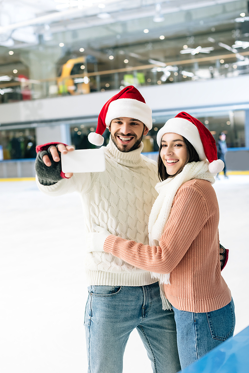 beautiful happy couple in santa hats taking selfie on smartphone on skating rink at christmastime