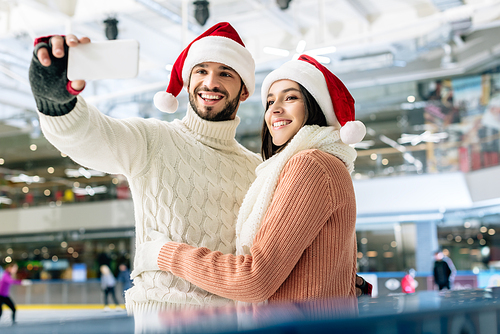 beautiful cheerful couple in santa hats taking selfie on smartphone on skating rink at christmastime