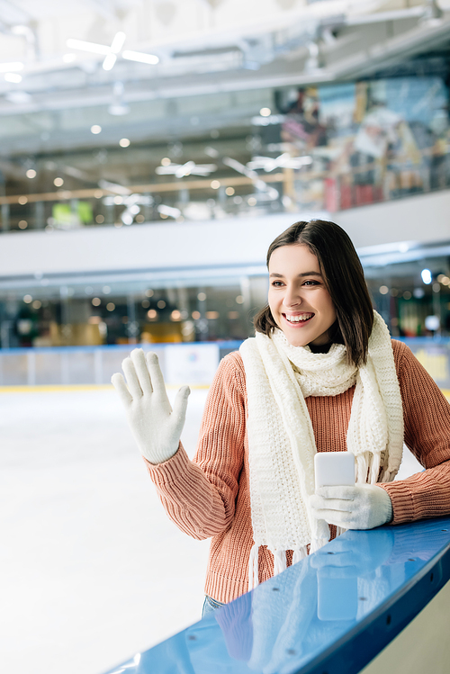 cheerful girl holding smartphone and waving hand on skating rink