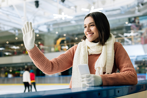 beautiful smiling girl holding smartphone and waving hand on skating rink