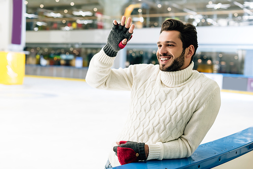 smiling man in sweater and gloves waving hand on skating rink