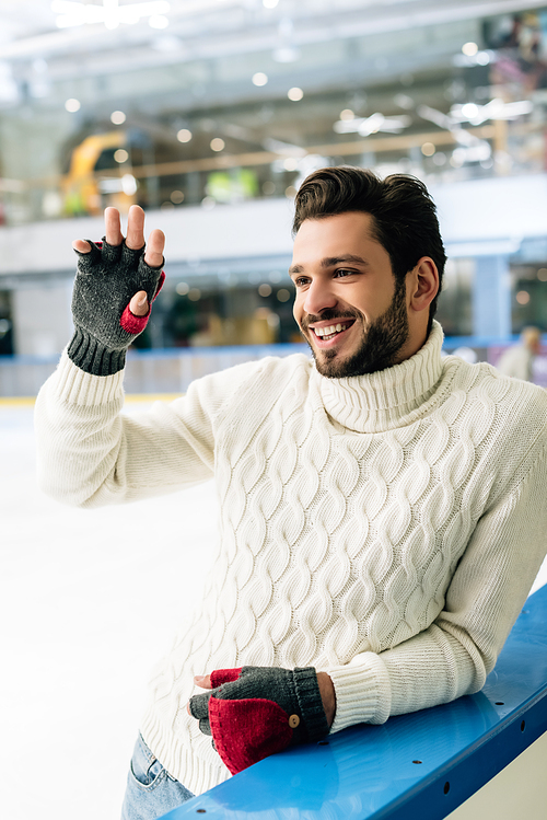 cheerful handsome man in sweater and gloves waving hand on skating rink