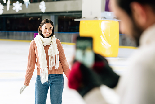 selective focus of boyfriend taking photo of smiling girlfriend on smartphone on skating rink