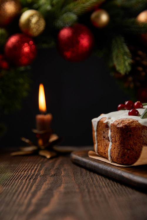 selective focus of traditional Christmas cake with cranberry near Christmas wreath with baubles and burning candle on wooden table isolated on black