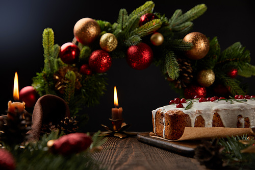 traditional Christmas cake with cranberry near Christmas wreath with baubles and burning candles on wooden table isolated on black