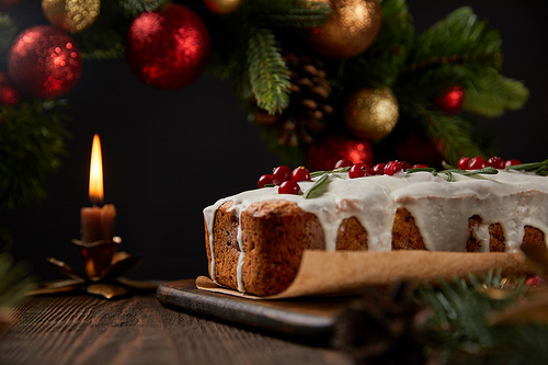 selective focus of traditional Christmas cake with cranberry near Christmas wreath with baubles and candle on wooden table isolated on black