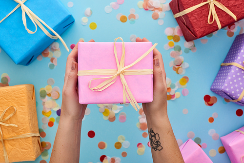 cropped view of woman holding wrapped pink gift box near colorful presents near confetti on blue