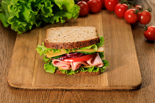 selective focus of fresh sandwich on wooden cutting board near lettuce and cherry tomatoes