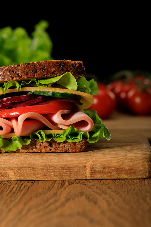 close up view of fresh sandwich with lettuce, ham, cheese, bacon and tomato on wooden cutting board isolated on black