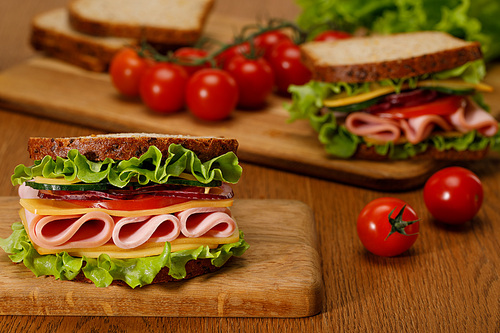 selective focus of fresh sandwich with lettuce, ham, cheese, bacon and tomato on wooden cutting board