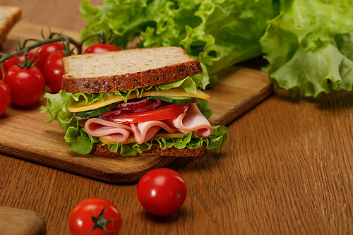 selective focus of sandwich on wooden cutting board near fresh cherry tomatoes and lettuce