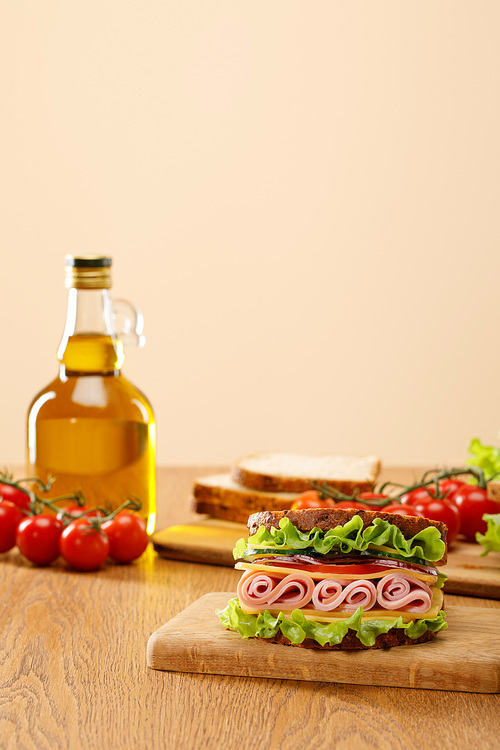 selective focus of fresh sandwich near lettuce, bread, cherry tomatoes and oil on wooden table isolated on beige