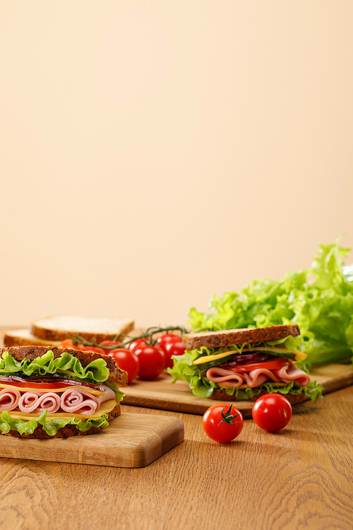selective focus of fresh sandwich near lettuce, bread, cherry tomatoes on wooden table isolated on beige