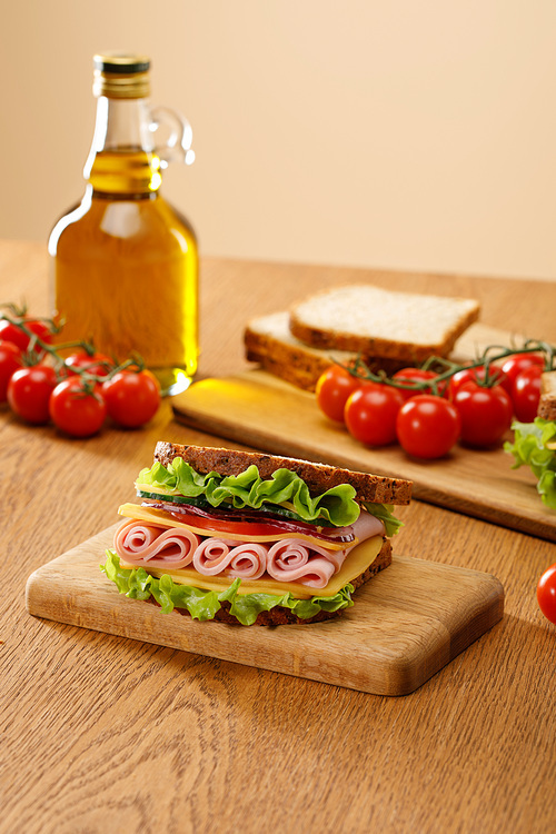 selective focus of fresh sandwich near lettuce, bread, cherry tomatoes and oil on wooden table isolated on beige