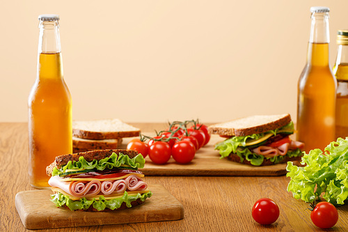 selective focus of fresh sandwich with lettuce, ham, cheese, bacon and tomato near bottles of beer at wooden table isolated on beige